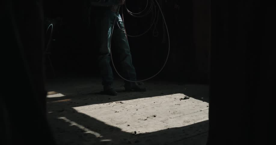 Person stands in a dark stable holding a lasso with light entering across the floor