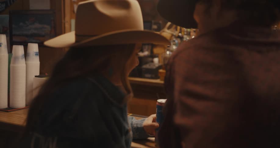 Woman wearing a cowboy hat talks at a bar counter with stacked cups in the background