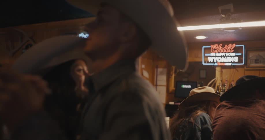 Young adult man and young adult woman wearing cowboy hats dancing together at a bar with other people talking and hanging around indoors during night time