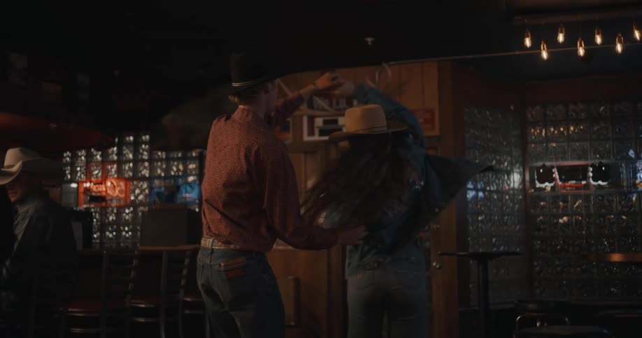 A man in a red shirt and a woman in a denim jacket dance together in a dimly lit bar performing spins and rhythmic steps on a wooden floor while other patrons sit at the counter in the background