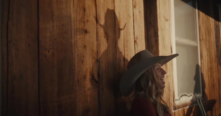 Woman wearing a cowboy hat leans against a sunlit wooden wall
