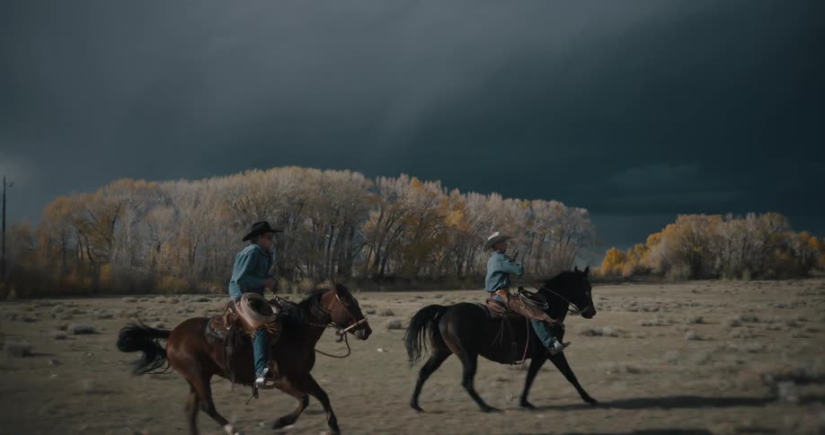Two adult men wearing cowboy hats riding horses on a field with very dark heavy clouds on the sky in the background outdoors