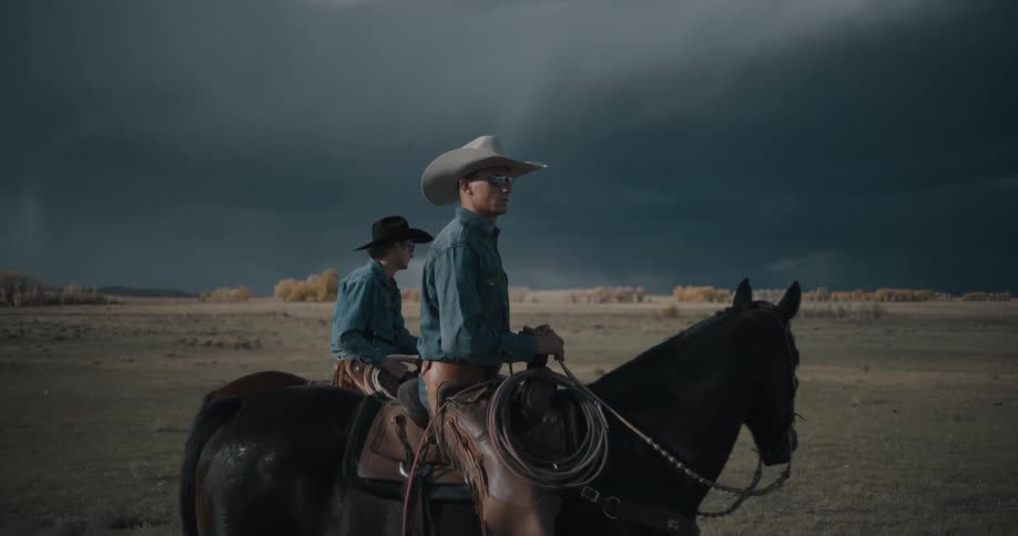 Two cowboys sit atop brown horses in a grassland