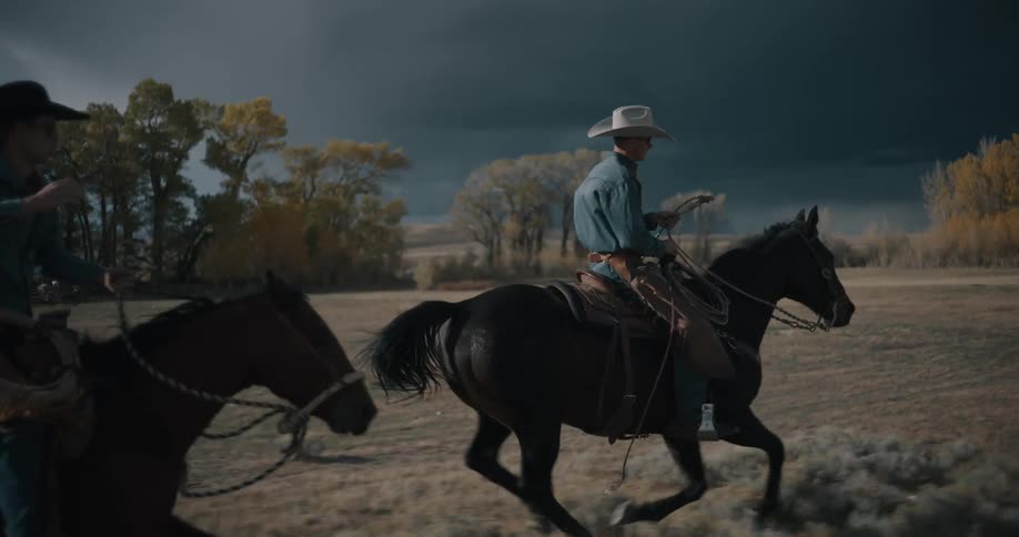 Two cowboys ride their horses across a vast open field under a dark stormy sky with one raising his hat in the air