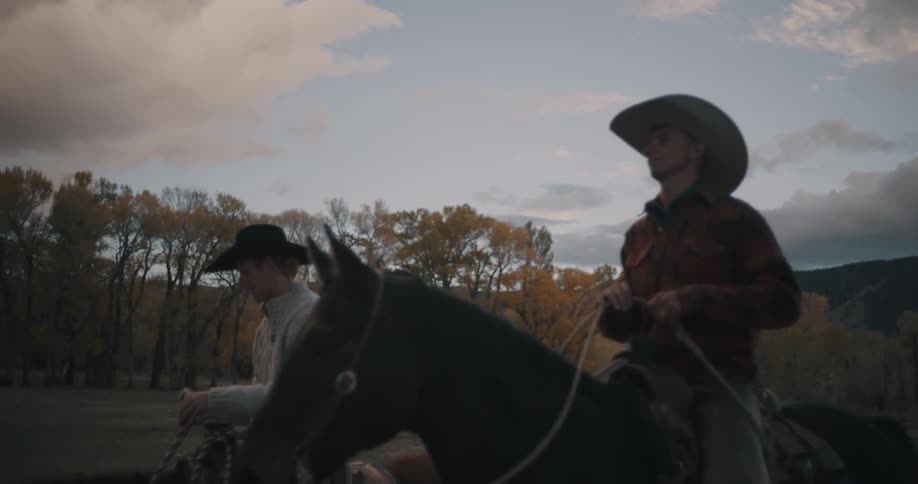 Two men wearing cowboy hats ride horses along a dirt path through a field at dusk with yellow autumn trees and large mountains visible under a cloudy sky in the background