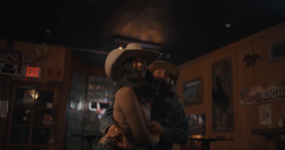 Women dance embracing each other in a bar wearing cowboy hats and with low lighting