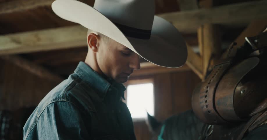 Young adult man wearing a cowboy hat and denim clothes saddling a horse at a stable indoors during day time