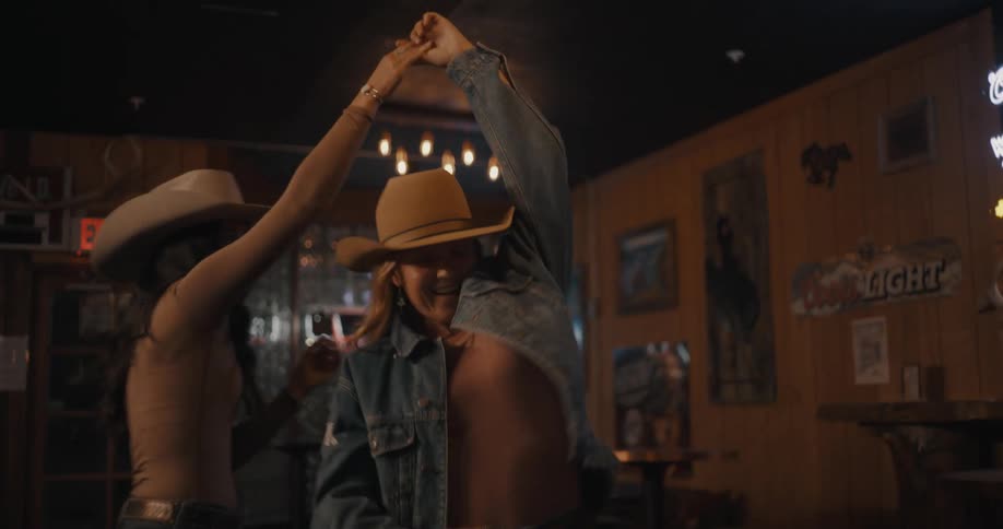 Two women dance in a bar wearing cowboy hats under low lighting