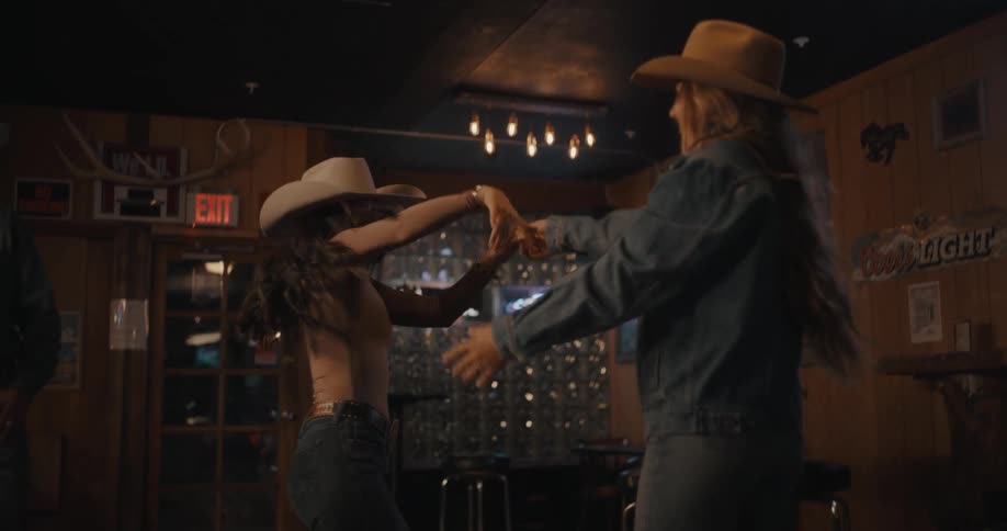 Two women dancing in a westernstyle hall wearing hats under low lighting