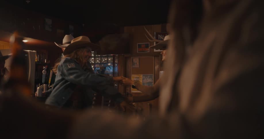 Two young women dancing in a pub while wearing cowboy hats with a man at the bar also in a cowboy hat watching the scene