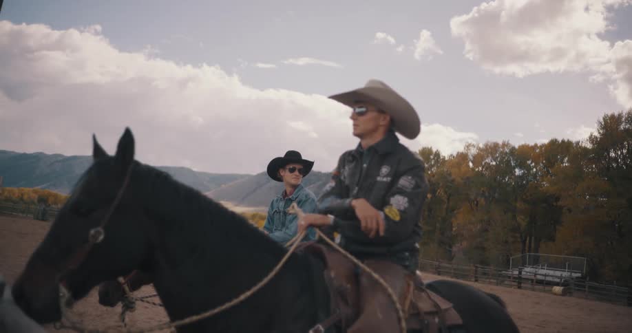 Two riders sit on horses in a rural area with mountains and trees in the background