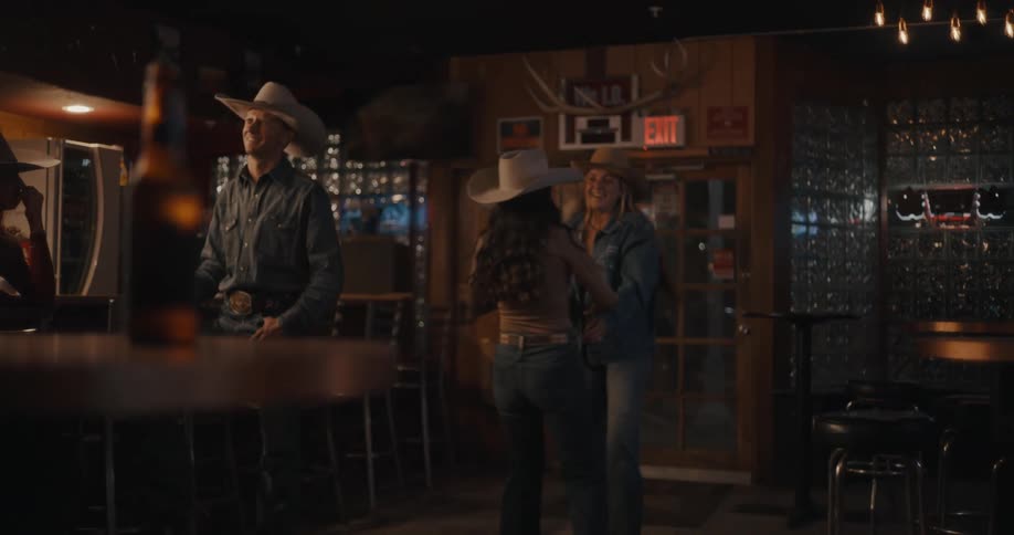 Two women dancing in a westernstyle bar wearing cowboy hats with a bar counter in the background