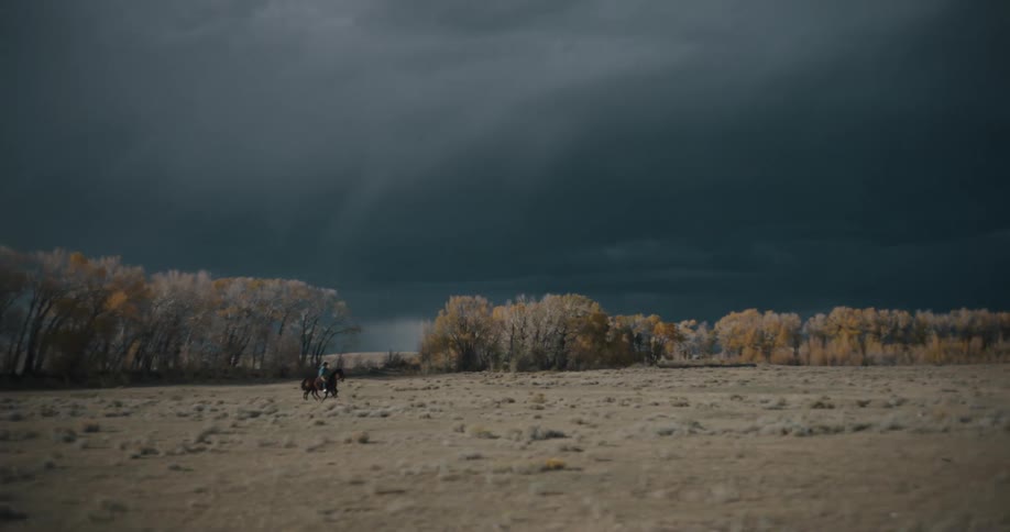 Riders cross an open field under a dark sky with trees in the background