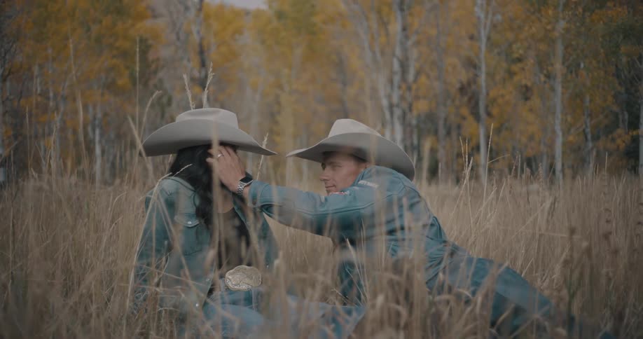 Couple sitting in a field wearing cowboy hats among tall grass with trees in the background