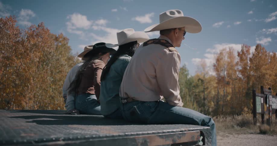 Four people sitting on the open tailgate of a truck all wearing cowboy hats with a man and woman seated together as a couple looking out at the wild trees on a sunny day