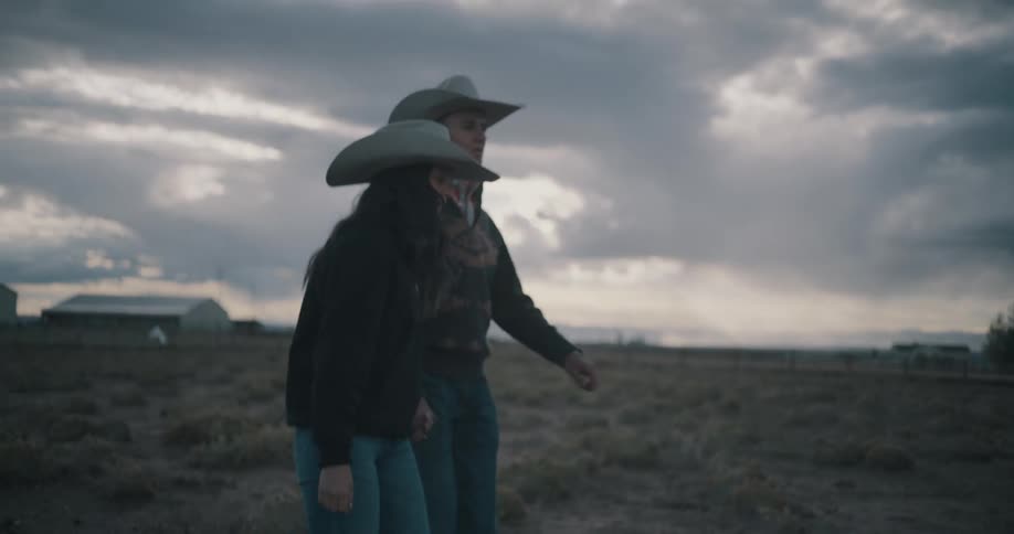 A couple wearing cowboy hats holding hands as they walk across a grassy countryside landscape on a cloudy day