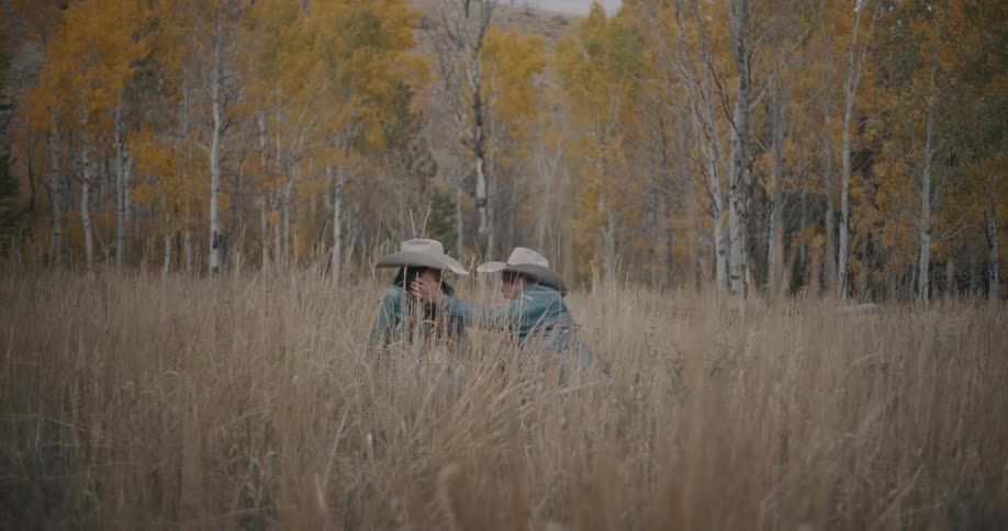 Couple sitting in a field of tall grass wearing cowboy hats with trees in the background