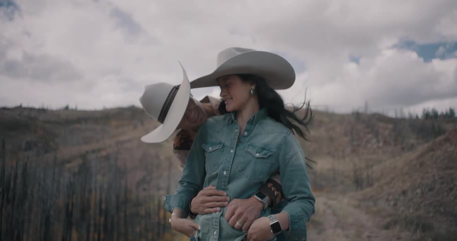 A man wearing a cowboy hat embraces a woman from behind as they stand together in a mountain landscape with dry brush and a cloudy sky