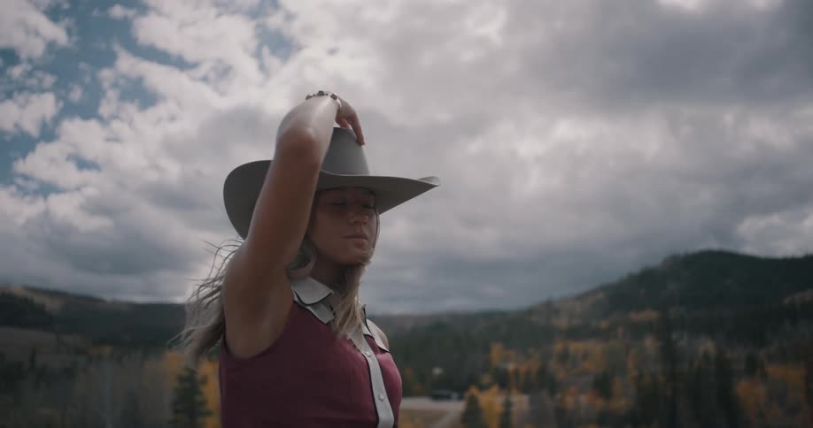 Woman wearing a hat holds the brim while looking over a mountainous landscape under a cloudy sky