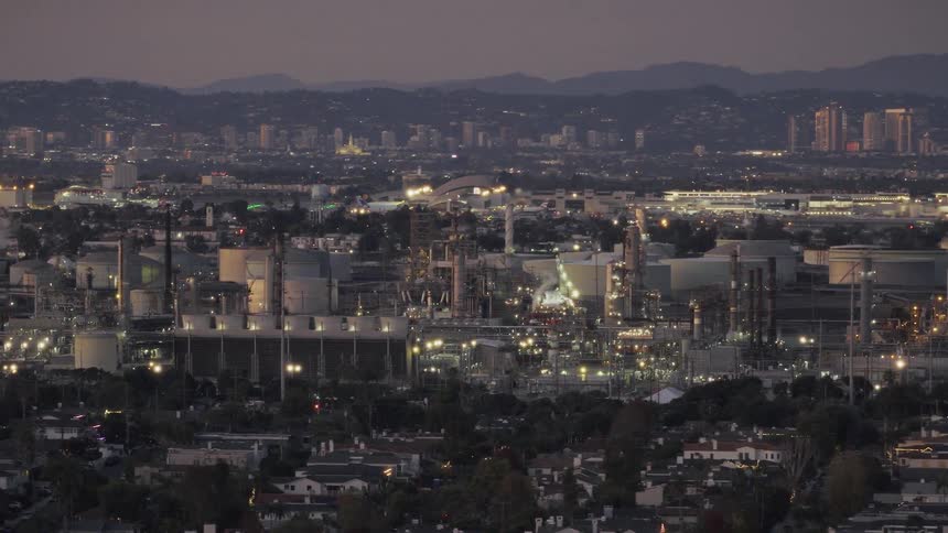 An industrial refinery with glowing lights and large storage tanks stands in the foreground of an expansive urban city landscape at dusk with a distant skyline and mountains visible under a dark evening sky