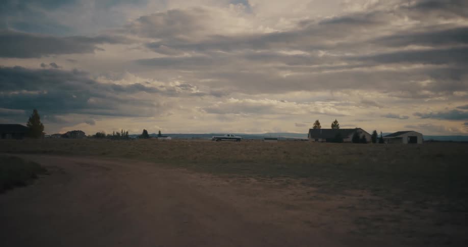Dirt road crossing an open rural area with distant houses under a cloudy sky