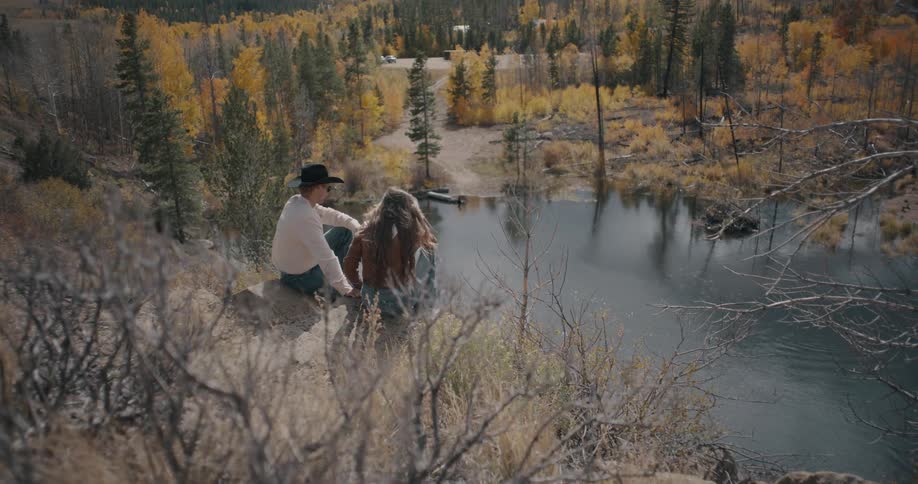 A man in a black hat and a woman with long hair sit on a rocky ledge overlooking a calm river and autumn trees