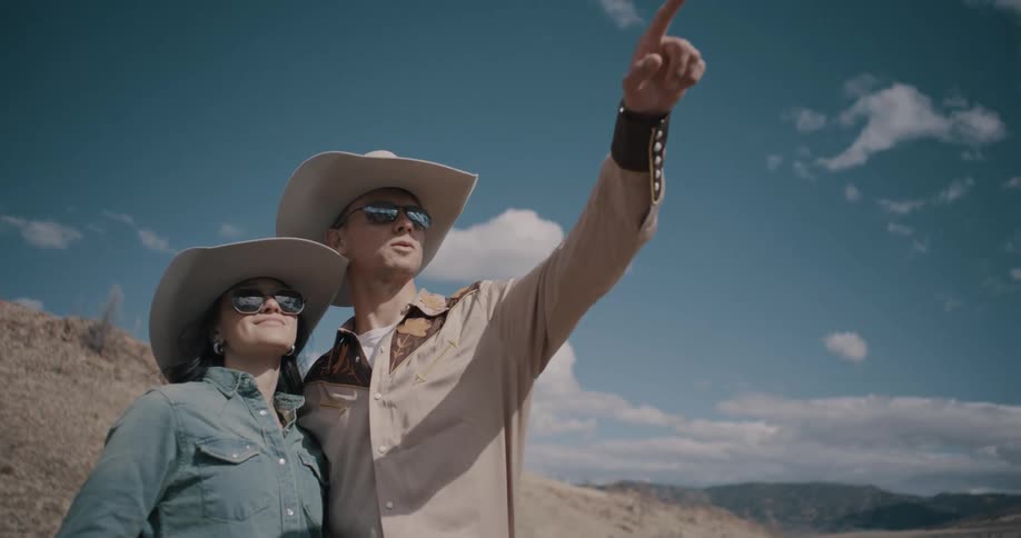 Couple wearing cowboy hats looks at the landscape while the man points toward the sky