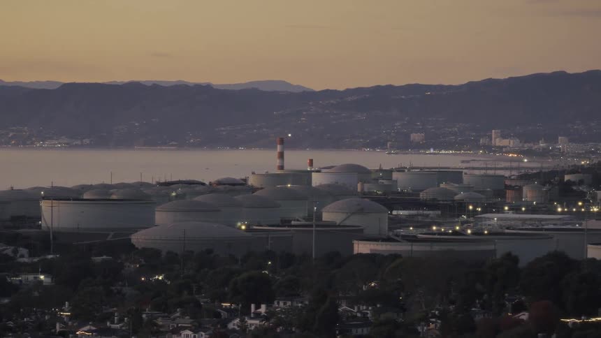 As dusk settles a sprawling coastal refinery sits against a backdrop of mountains and water its large storage tanks dotting the industrial landscape