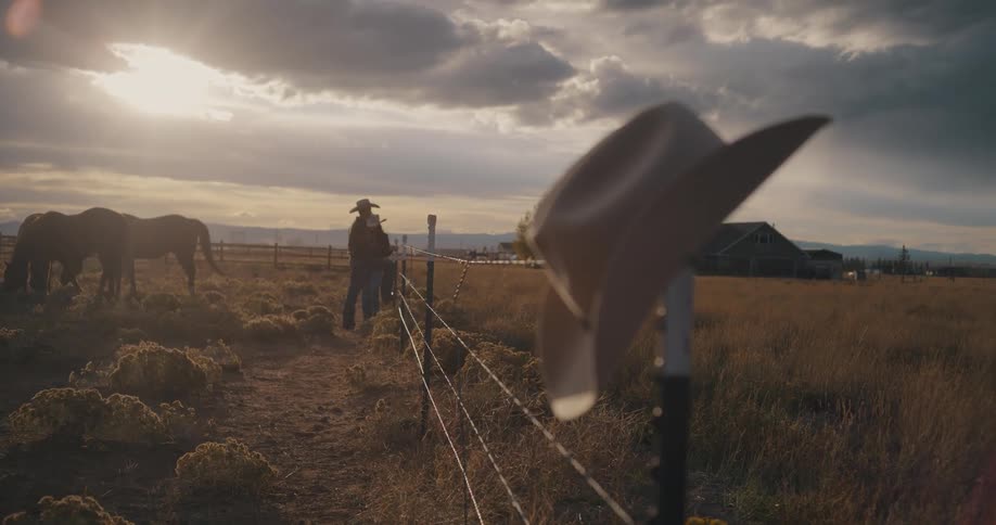 Cowboy hat hanging on a fence while a person stands in the background in a rural area at sunset