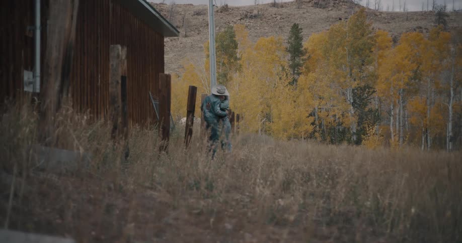 A man and a woman wearing denim and cowboy hats walk through tall grass past a wooden building and embrace under a grove of yellow autumn trees