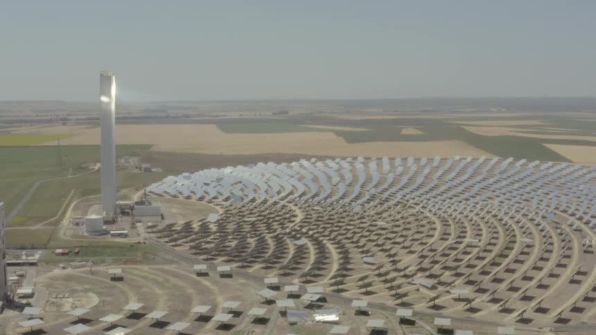 An aerial view slowly orbits a massive solar power plant where hundreds of gleaming mirrors reflect sunlight onto a tall central tower
