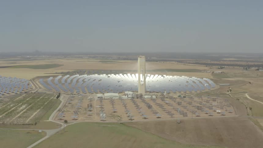 From high above the view slowly pulls back to reveal a massive solar power plant its central tower encircled by a vast array of gleaming mirrors spread across the arid landscape