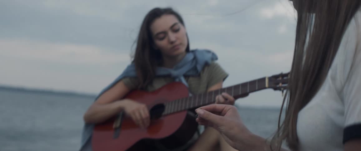 Two women sit on the beach while one plays guitar