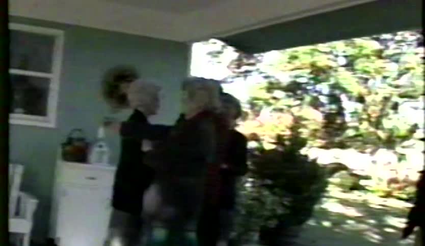 Vintage home video footage shows people in formal clothing walking across a lawn and gathering on a house porch to socialize on a sunny day