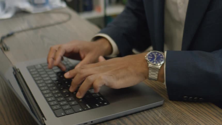 Hands type on a laptop placed on a desk with a metal wristwatch visible on the wrist