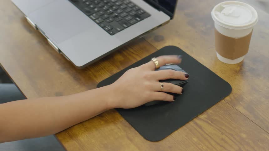 A woman with rings and dark nail polish moves a black mouse on a pad while working at a wooden desk with a laptop and a coffee cup