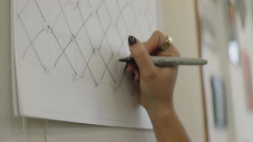 Closeup of a hand using a black marker to cross off dates on a paper wall calendar