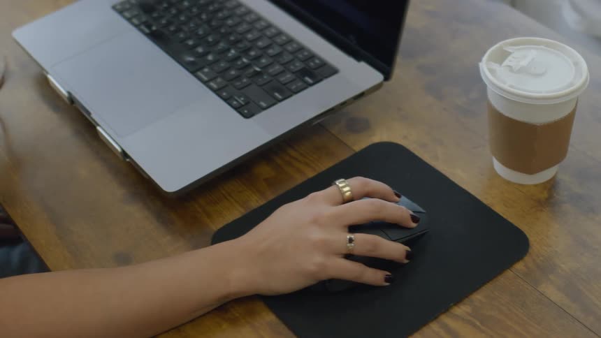 A woman with black nail polish and gold rings uses a wireless mouse on a black pad next to a laptop and a coffee cup on a wooden desk