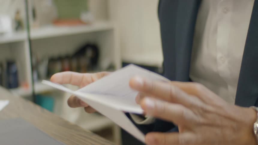 A man wearing a blue suit and a silver watch opens a white envelope and removes a paper document to read it while sitting at a desk with a laptop