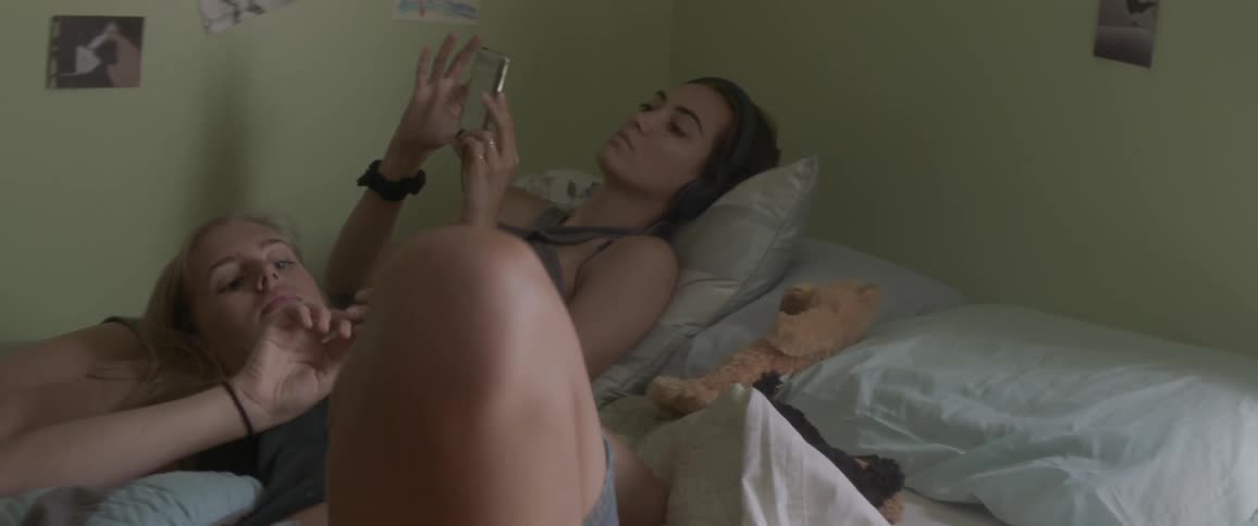 Woman listening to music while another woman rests head on her legs in bed
