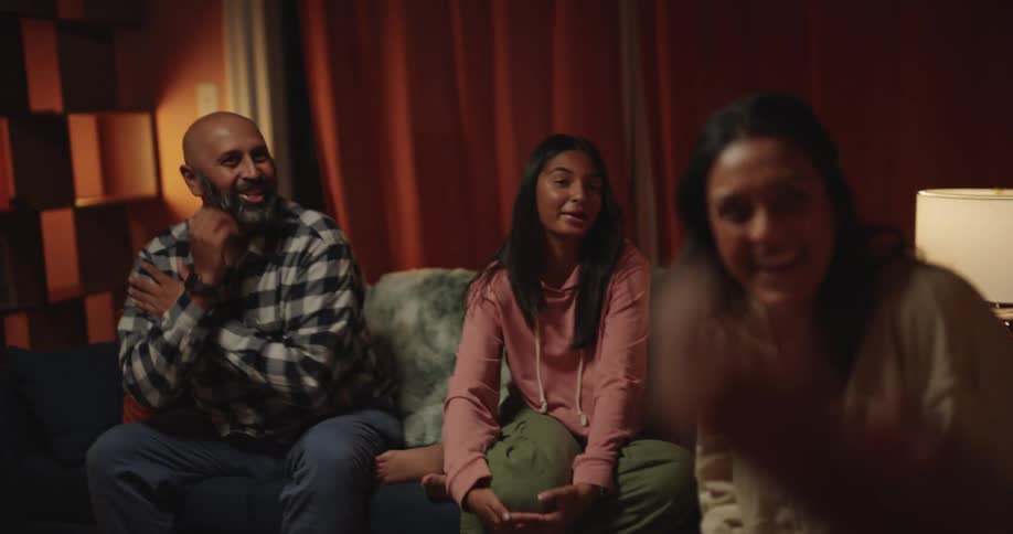 A man with a beard a woman and a teenage girl sit on a sofa and laugh while watching a young girl perform karate poses in a living room with warm lighting during the evening