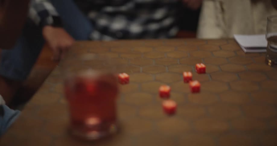 Teenager boy shaking dice on a cup and rolling them over a wooden coffee table with an adult man and a woman sitting next to it on the floor indoors