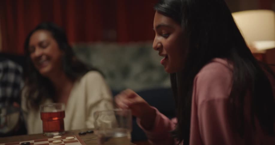 A young woman focuses on a game of checkers successfully jumping her piece over another on the board she celebrates her move with a joyful expression while being cheered on by others in the background