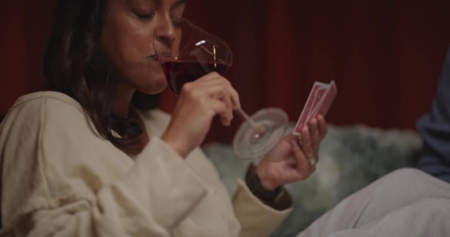 Young adult woman drinking wine and holding playing cards on her hand sitting on a couch with a man next to a coffee table with another glass and a wooden board with cheese and grapes over it indoors