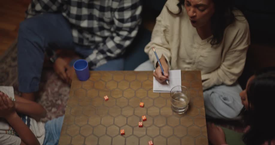 A man shakes and rolls several red dice from a blue cup onto a hexagonalpatterned table while a woman next to him writes on a notepad two young girls sit around the table and watch the dice land as the family engages in a game night together indoors