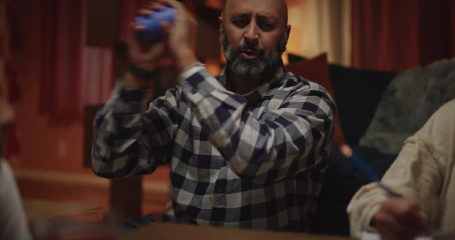 A man with a gray beard wearing a plaid shirt shakes a blue cup and celebrates with raised arms while playing a board game with a woman and two girls in a room with warm lighting