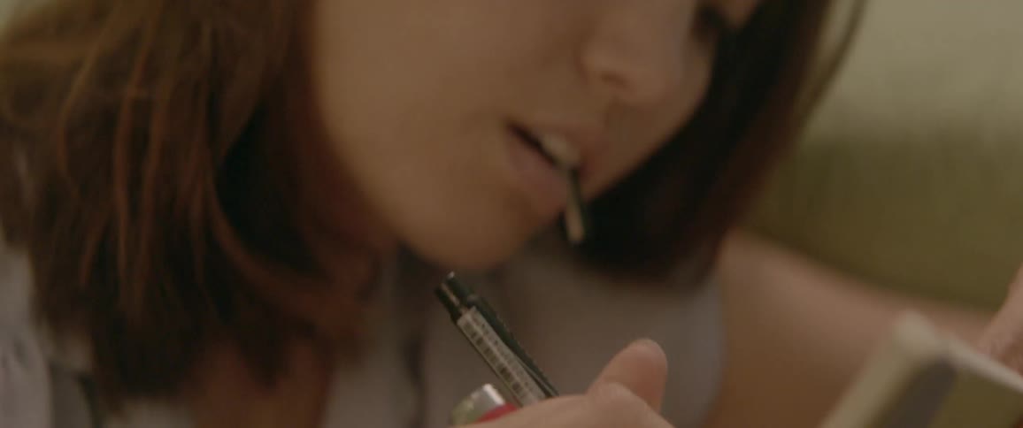 A woman holds a pen cap in her teeth and uses the pen to write in a book while holding a lighter