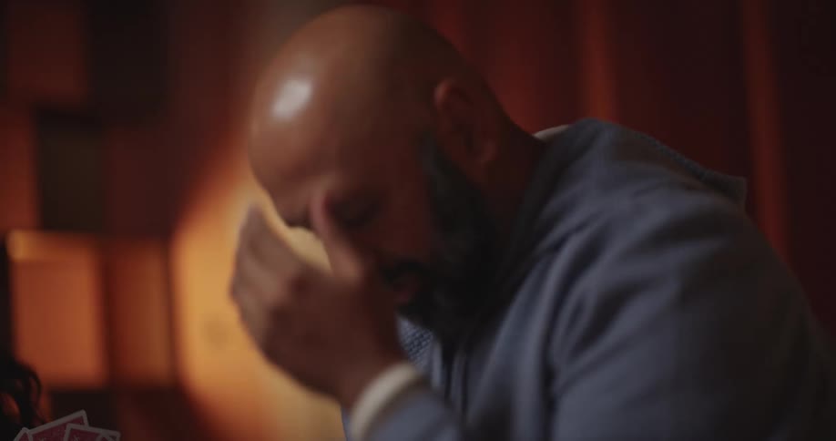 A woman holding a hand of playing cards sits near a man with a beard who reacts with disappointment while wine and grapes rest on a table in a dimly lit room with warm orange tones