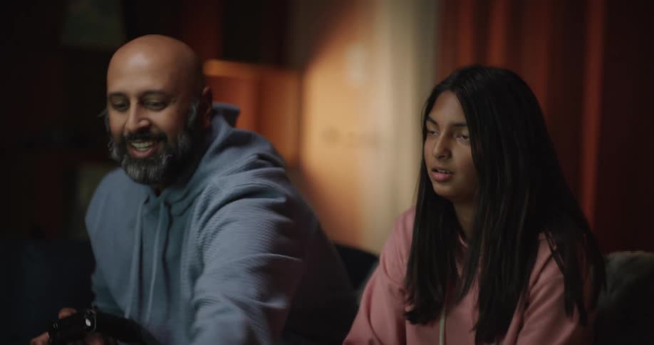 A father and daughter sit together on a couch focused on playing a video game with handheld controllers they react with excitement and surprise to the action on the screen sharing a bonding moment in their dimly lit living room