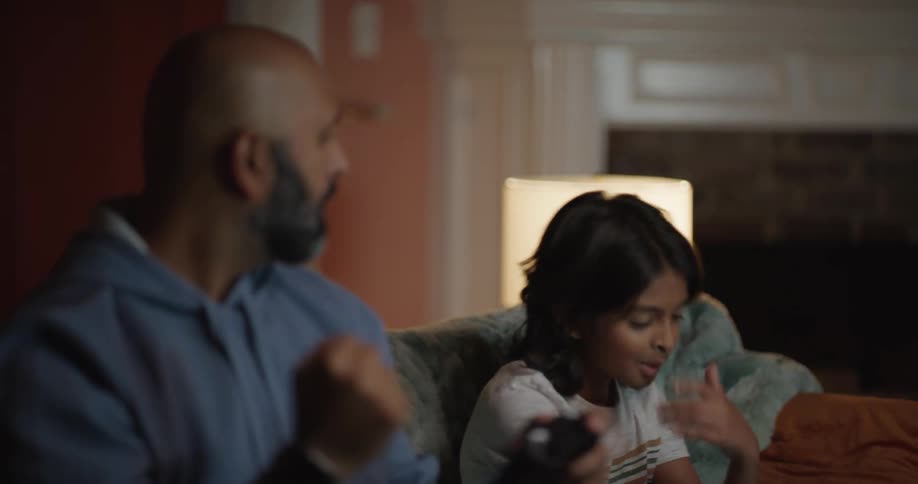 A man with a gray beard and a young girl sit on a sofa in a dimly lit living room while the girl holds a game controller and reacts with disappointment to a screen